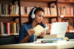 A student wearing headphones holds a book and looks at a laptop in a library.